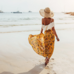 Donna che cammina lungo la spiaggia con un cappello di paglia e una gonna gialla a fantasia floreale ed un top bianco