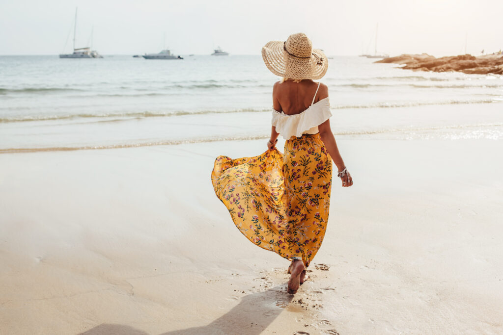 Donna che cammina lungo la spiaggia con un cappello di paglia e una gonna gialla a fantasia floreale ed un top bianco