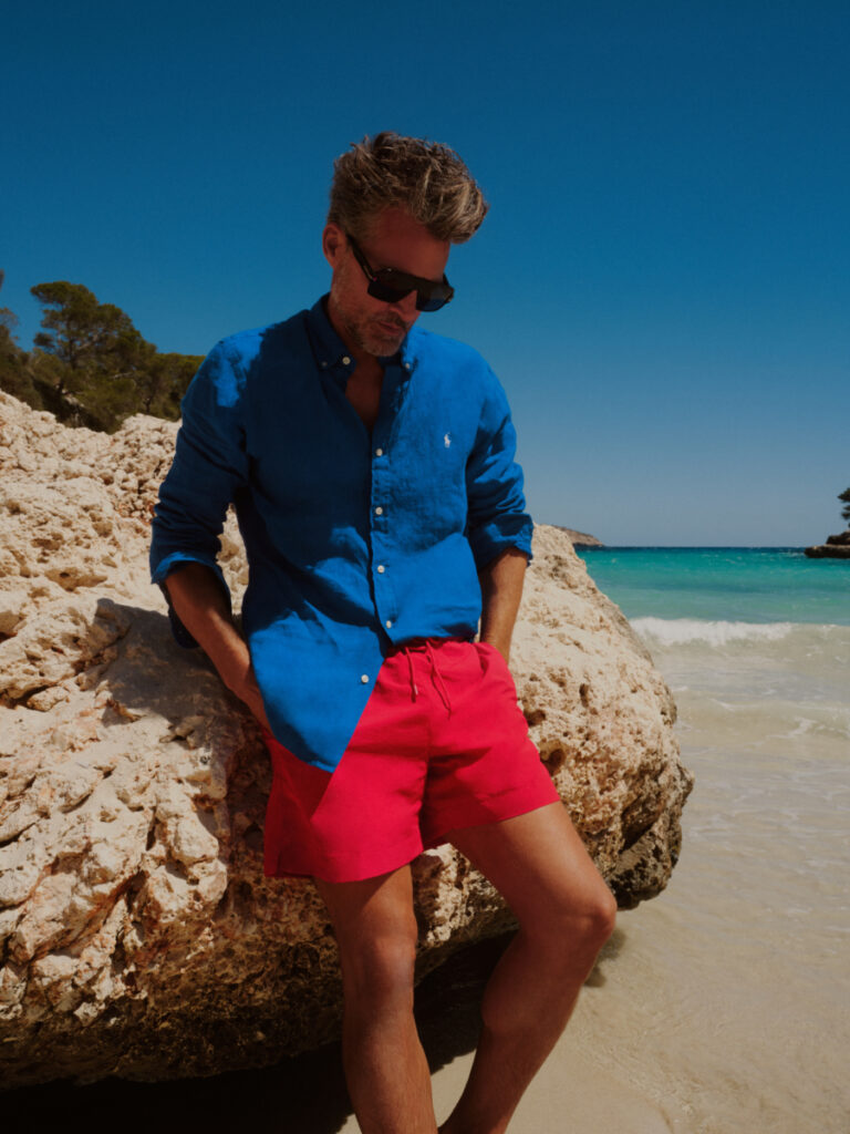 uomo in spiaggia con pantaloncini da mare rossi, camicia blu e occhiali da sole